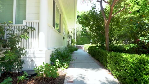 Walking along a garden path beside a white wooden porch on a sunny day Stock Footage 345679013