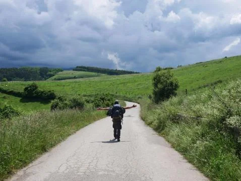 Walking man with backpack surrounded by green hill prairie with trees and clo Stock Photos