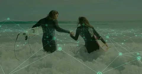 Walking pair in wetsuits holding hands, wading at shore with pale-pink board, Fotos Stock