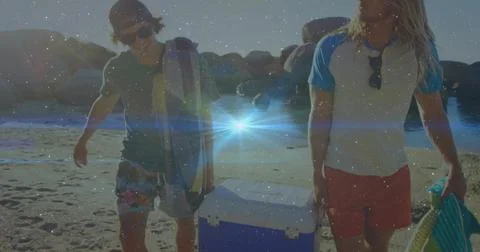 Walking two men carrying blue cooler, board on low-tide beach, wearing backward Stock Photos