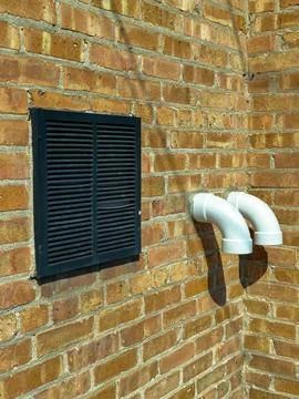 A wall made of red bricks shows a black vent cover and two white exhaust pipe Фото