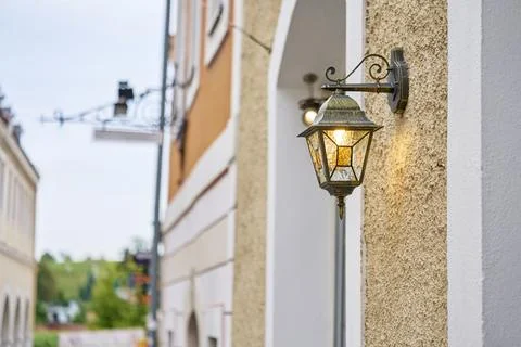 Wall mounted lantern glowing on textured building facade beside narrow street Stock Photos
