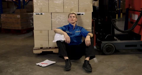 Warehouse engineer in blue uniform sits on floor beside cardboard boxes with Stock Footage 307008131