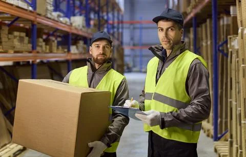 Warehouse staff checking inventory clipboard while coworker carries box in aisle Stock Photos