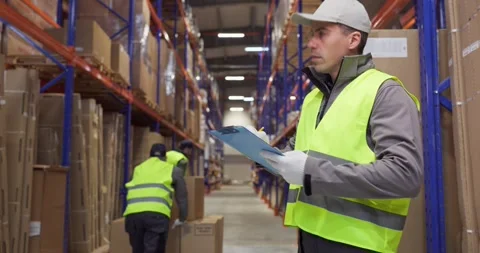 Warehouse worker checks inventory with clipboard among tall shelves and boxes Stock Footage 335602742