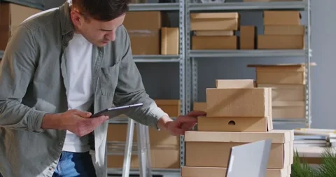 Warehouse worker looking at stack of packages, using tablet and entering data in Stock Footage 277643685