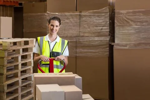 Warehouse worker operating pallet jack loaded with boxes among pallet stacks in Stock Photos