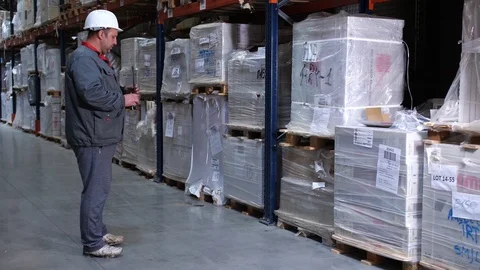 Warehouse worker rewrites the goods. A man in a helmet and a uniform with a Vídeo Stock 111766137