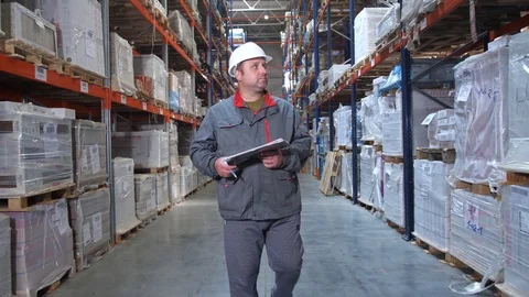 A warehouse worker in uniform walks along the racks, he holds a folder in his Stock-Footage 111766428