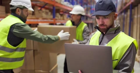 Warehouse worker using laptop for inventory as colleagues move cardboard boxes Stock Footage 327357031