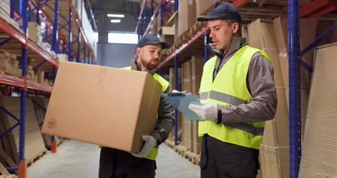 Warehouse workers carry box while colleague checks clipboard in logistics aisle Stock Footage 331074286
