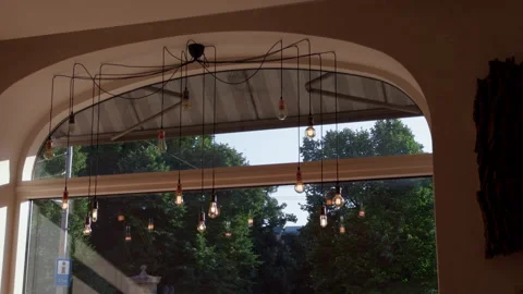 Warm cafe interior with arched window, hanging bulbs, and leafy trees outside in Stock Footage 344975349