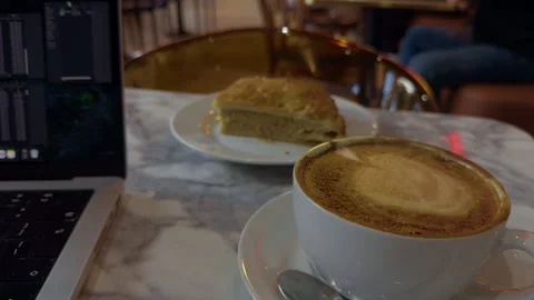 Warm cafe table with cappuccino, cake slice, and laptop beside marble surface Stock Footage 344966333