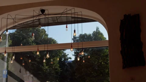 Warm rustic cafe interior with hanging bulbs, arched window, and leafy view Stock Footage 345454149