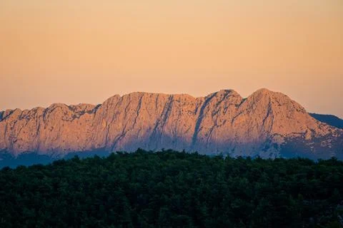 Warm sunset light covers rugged mountain ridge above dark forest Stock Photos
