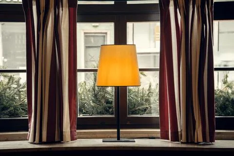 A warmly lit table lamp on a rustic windowsill, surrounded by striped curtains Foto stock