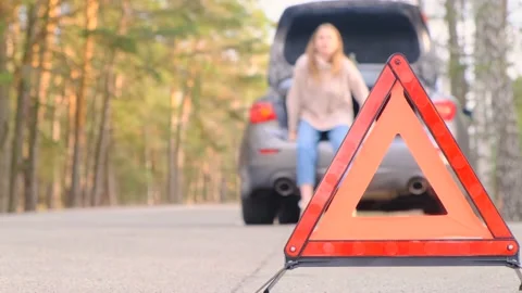 Warning red triangle. Sad young woman sits in trunk of gray car with flashing 스톡 동영상 163997346