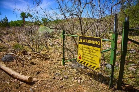 Warning Sign on Barbed Wire Gate in Rugged Desert Landscape Eye Level View 스톡 사진