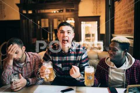 Watching TV in bar. Three happy young men drinking beer and gesturing with di Stock Photos