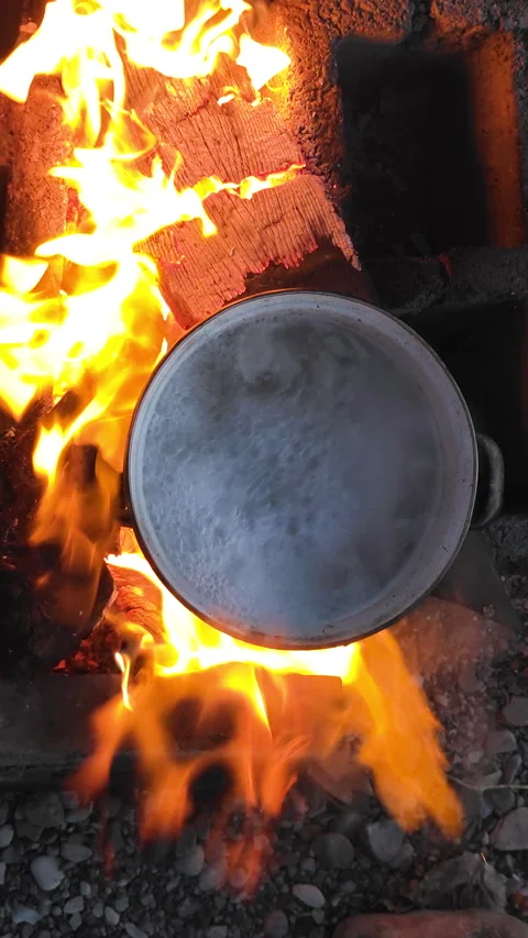 Water boiling in a pot over a campfire on a pebble beach with bright flames and 스톡 동영상 329619397