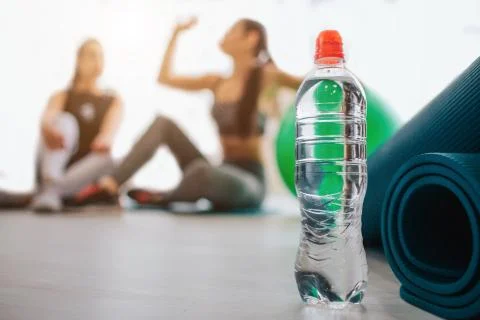 Water bottle stand in at mat. Two young women sit behind. One of them drink Foto stock