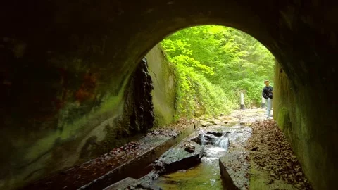 Water flowing through a dark concrete tunnel with a man standing at the exit Stock Footage 334868940