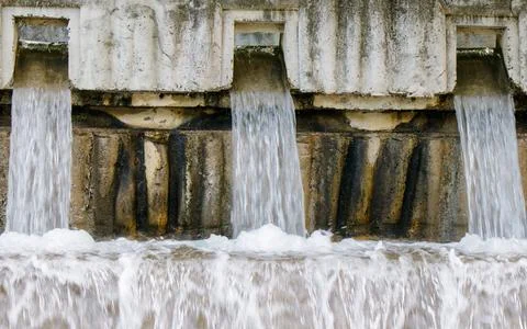 Water flows from concrete structure at a dam during daytime near a river Stock Photos