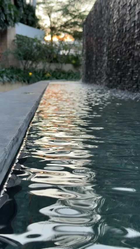 Water flows down a vertical fountain wall and falls into a pool while sunlight Stock Footage 330663183