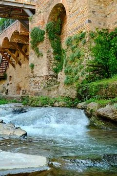 Water flows gently over rocks near a historic stone wall covered in greener.. Stock Photos