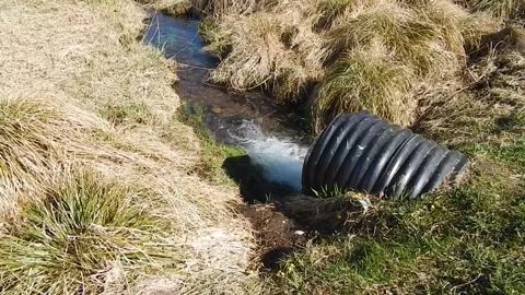Water flows out of a large black corrugated pipe into a river. Stock Footage 309000759