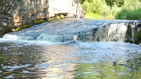 Water flows over a small waterfall in a forest setting near a river during dayti Stock-Footage 325625557