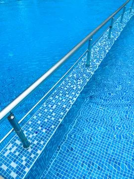 Water flows over a tiled pool edge with a metal railing in a bright blue in.. Stock Photos