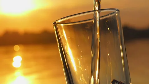 Water pours in glass in rays of setting sun on shore of lake Stock Footage 253903035