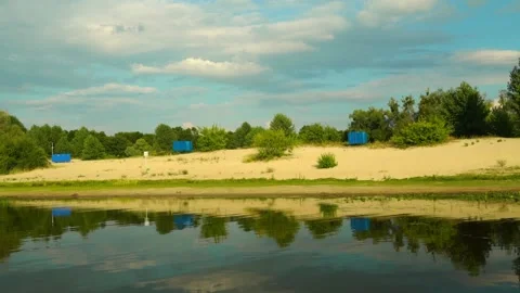 Water reflects trees and clouds by the sandy riverbank near blue containers in a Stockbeeldmateriaal 340448020