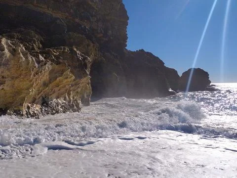 Water splashes against the rugged cliffs as waves roll in, sparkling under br Stock Photos