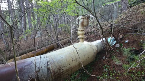 Water is vigorously leaking from an old, rusty industrial pipeline laying on the Stock Footage 335270821