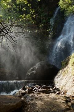 Waterfall and lower cascade in the forest with water fog lit with sunbeams Stock Photos