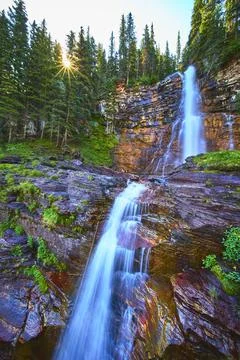 Waterfall Cascading Down Rocky Cliff with Sunburst Through Pine Forest 스톡 사진