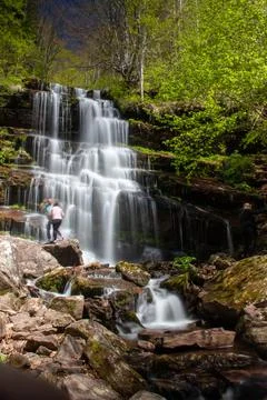Waterfall cascading down rocky hillside with two people exploring the lush .. Stock Photos