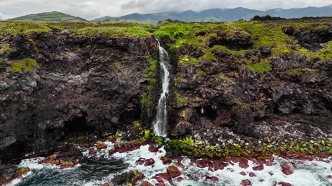 Waterfall dropping from coastal cliff into ocean with volcanic stones Stock Footage 340069996