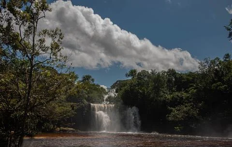 A waterfall falling into a gentle river next to a forest with white clouds .. Stock Photos