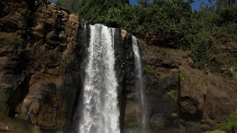 Waterfall Flowing Down Rocky Cliff, Lush Jungle, Clear Blue Sky, Tilt Down Shot Stock Footage 324771273