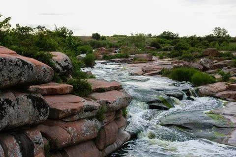 Waterfall flows among high rocks, red stones and cliffs, green bushes and tre Stock Photos