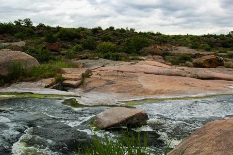 Waterfall flows among high rocks, red stones and cliffs, green bushes and tre Stock Photos