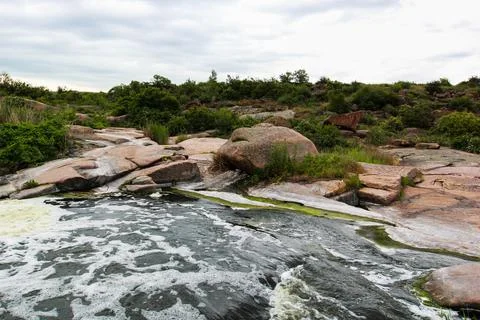 Waterfall flows among high rocks, red stones and cliffs, green bushes and tre Stock Photos