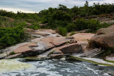 Waterfall flows among high rocks, red stones and cliffs, green bushes and tre Stock Photos