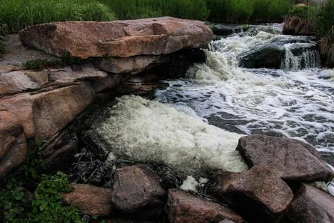 Waterfall flows among high rocks, red stones and cliffs, green bushes and tre Stock Photos