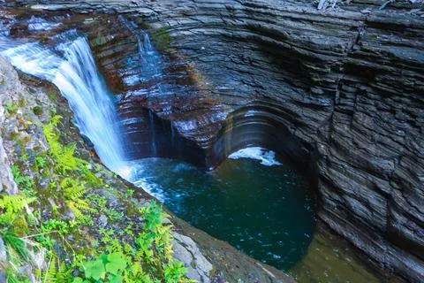 The waterfall flows into a moss-covered pool surrounded by rocks and plants.. 스톡 사진