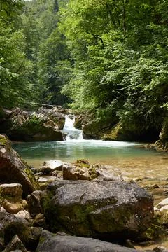 The waterfall flows into mountain lake in forest, large stones by stream, clear Stock Photos