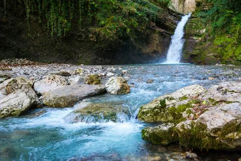 Waterfall in a green spring forest surrounded by stones, clear turquoise wate Stock Photos